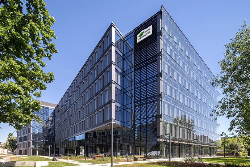 Natural Pharmaceuticals Hepactum&trade; FORTE Kaasaegne klaasfassaadiga büroohoone roheliste puudega linna rajoonis.